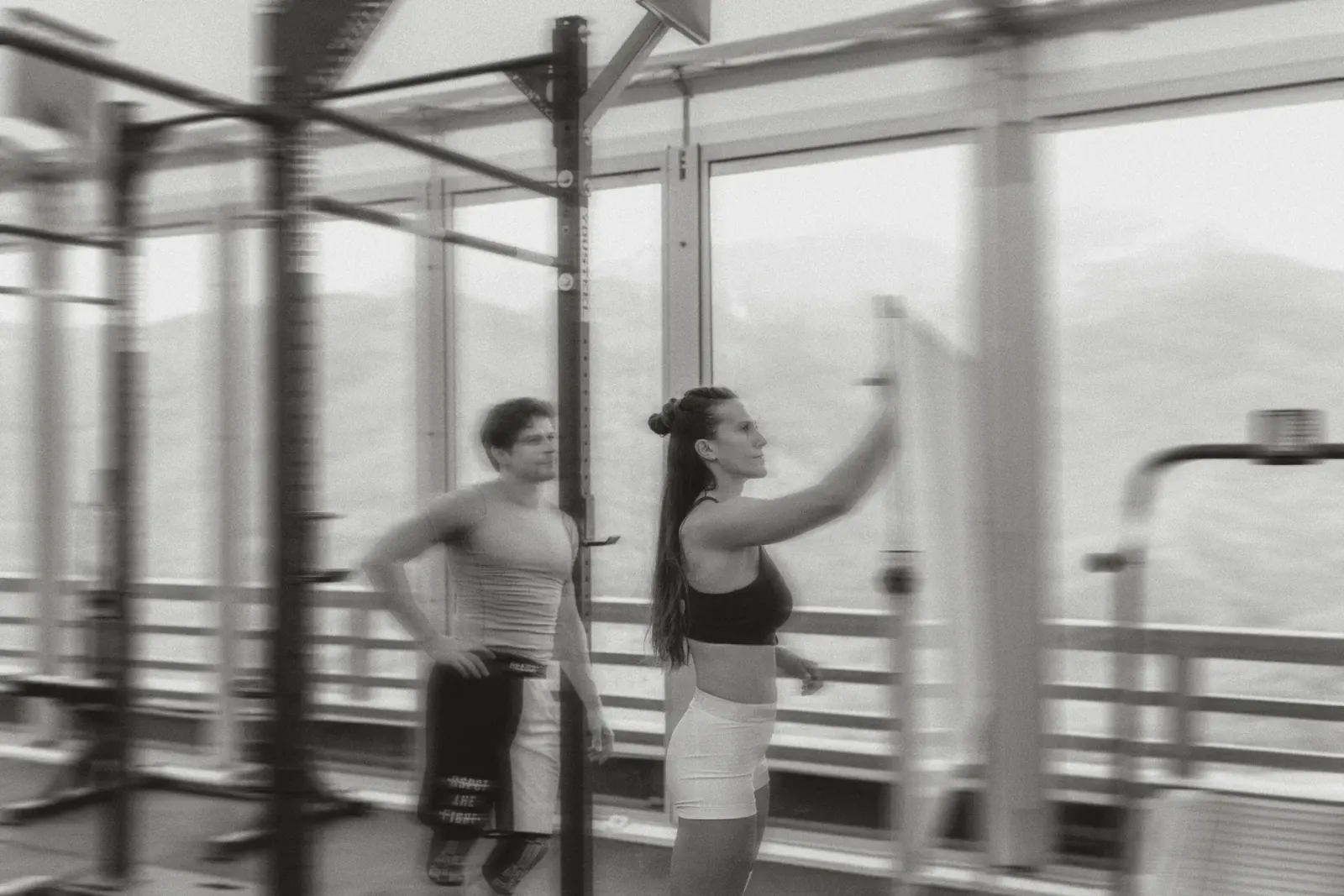 Dos personas entrenando en un gimnasio con racks y ventanales