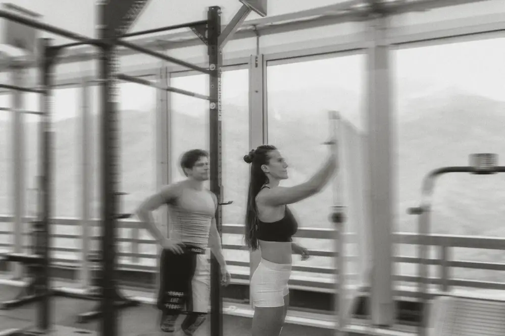 Two people training in a gym with racks and large windows